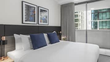 Quarto de hotel moderno com uma cama grande, mesa de cabeceira, abajur e janela com vista para prédios.
