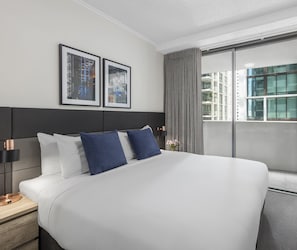 Ein modernes Hotelzimmer mit einem großen Bett, einem Nachttisch, einer Lampe und einem Fenster mit Blick auf Gebäude.