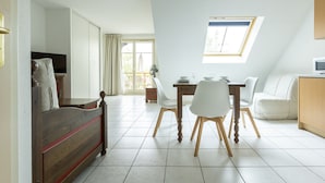 A modern living room with a dining area, a sofa, a TV, and a kitchen area with a window. - Résidence Odalys Le Domaine des Rois (Bergheim)