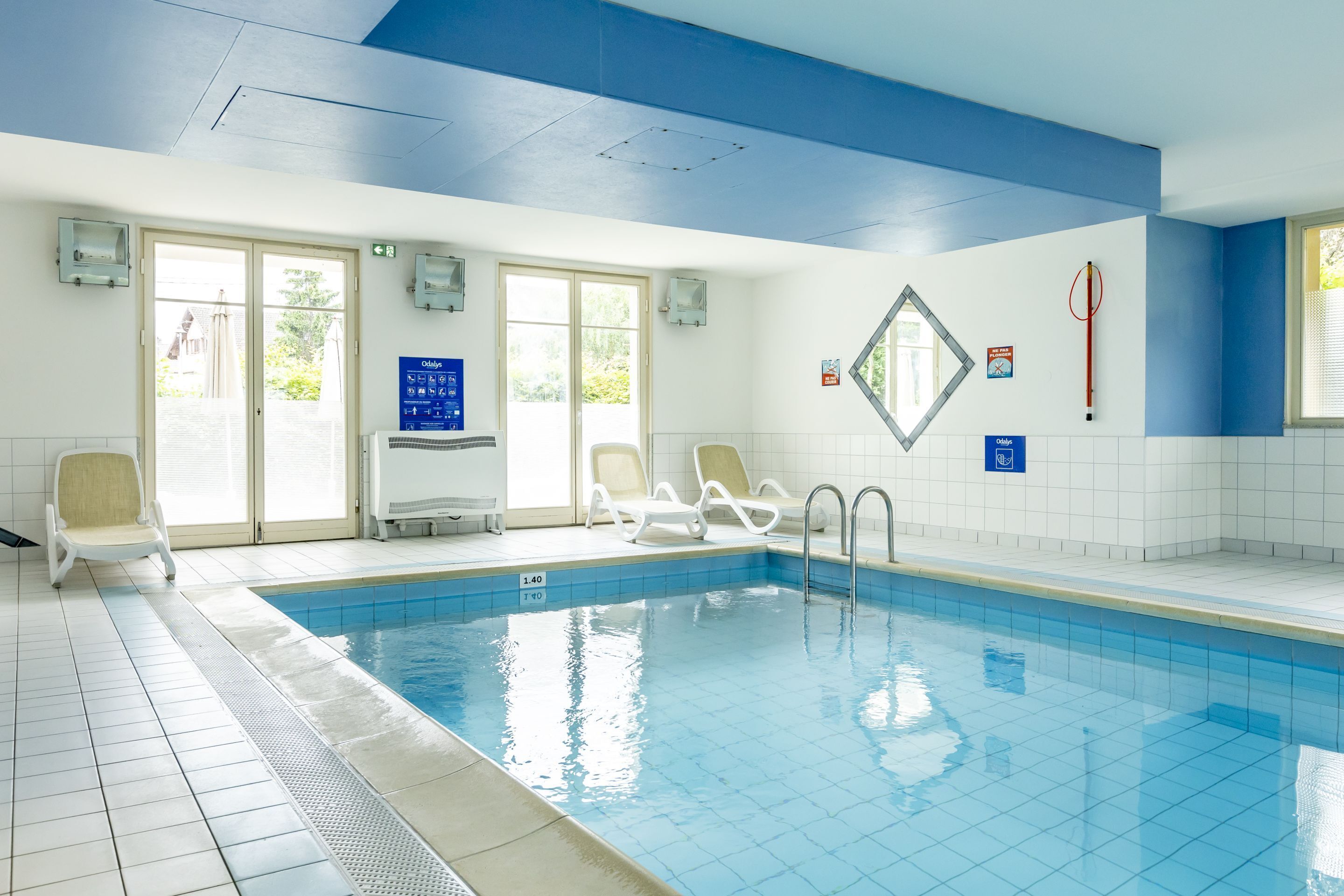 A swimming pool with clear blue water, white tiled walls, and a blue ceiling. There are two lounge chairs and a window with blinds.