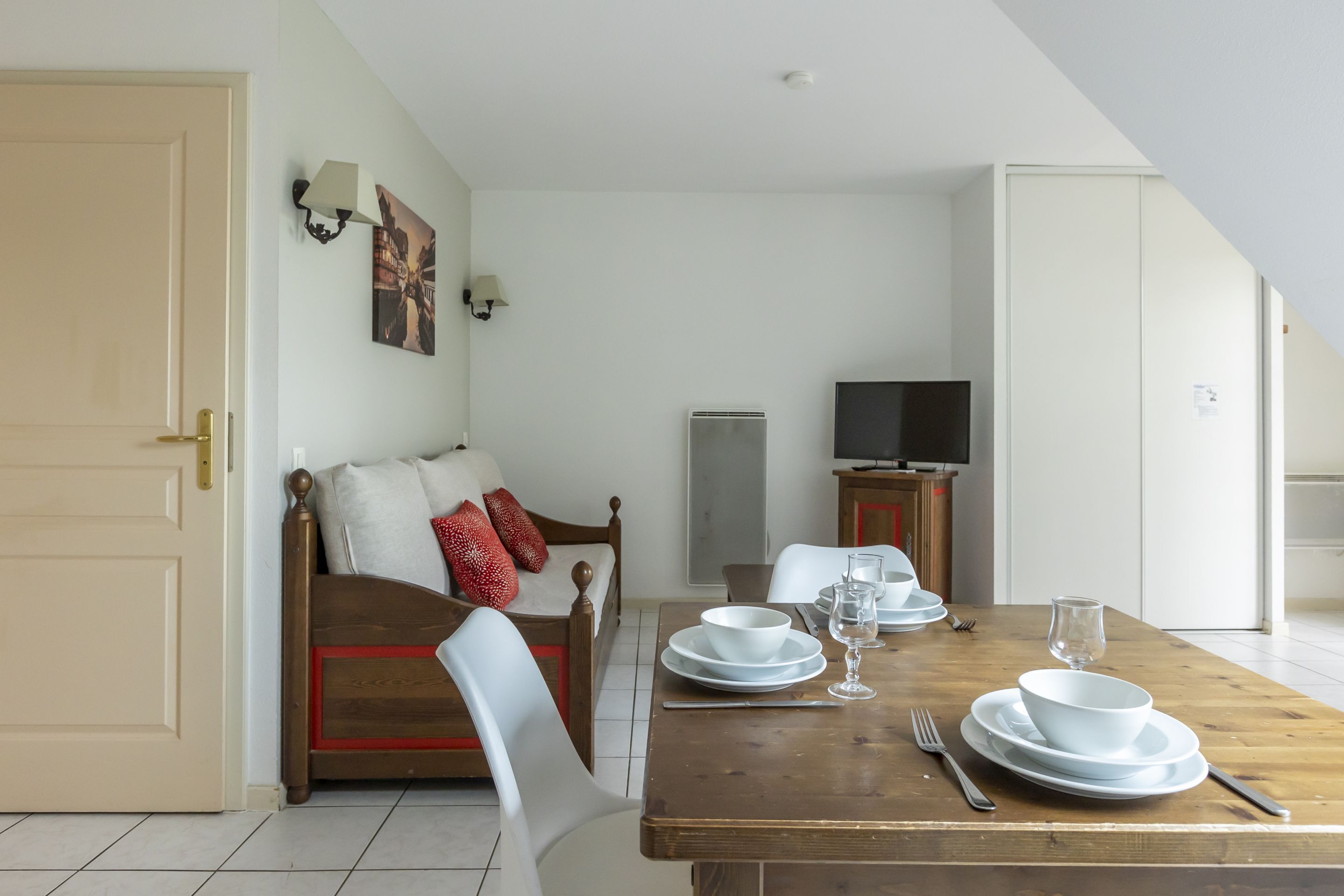 A dining area with a wooden table set for two, a sofa with red pillows, a television, and a white door.