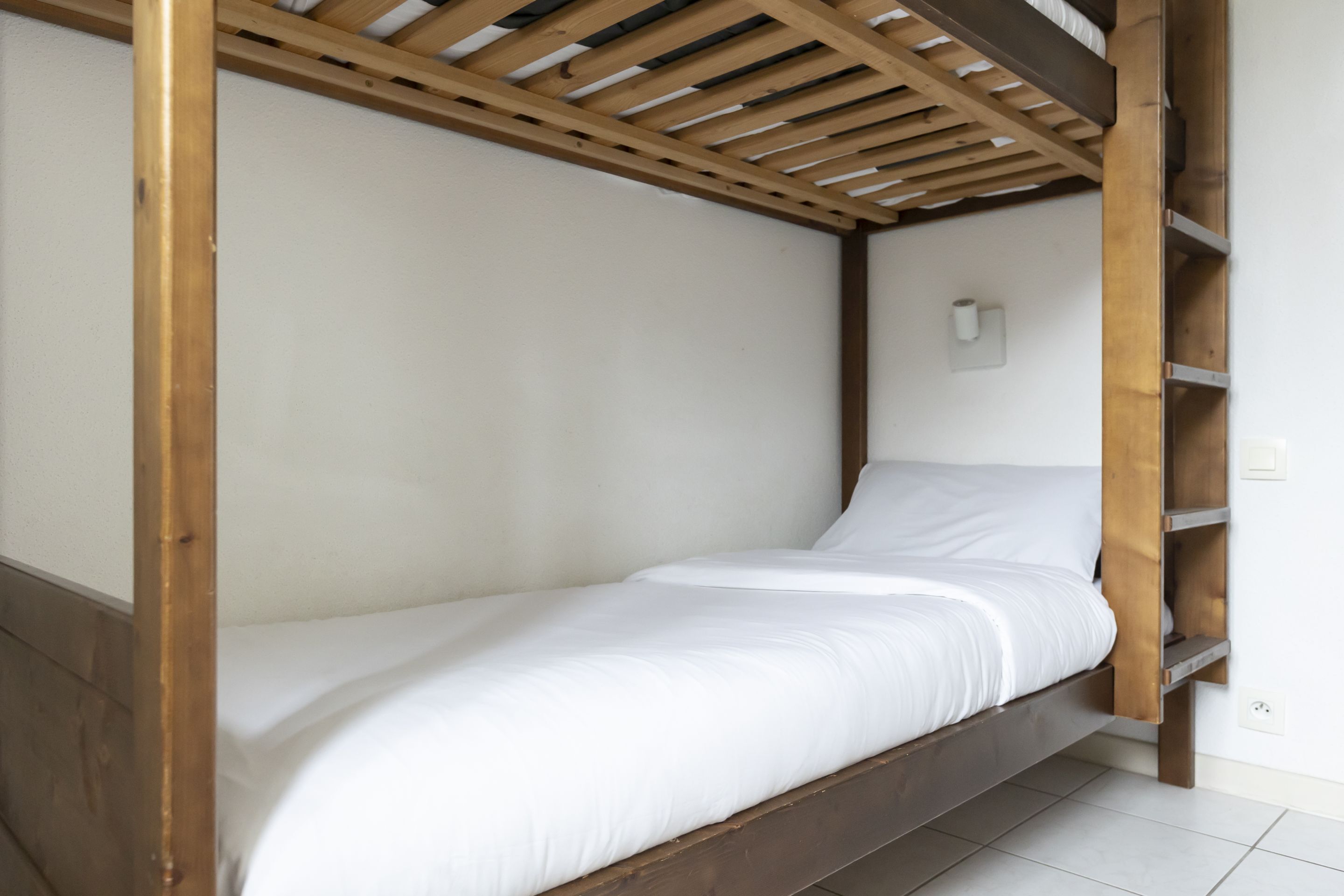 A single bed with a wooden frame and white bedding in a room with white walls and a tiled floor.