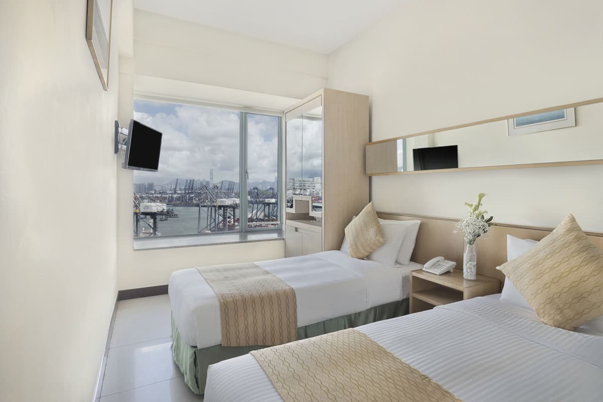 Une chambre d’hôtel avec deux lits, une grande fenêtre donnant sur la ville, un téléviseur à écran plat fixé au mur et une table de chevet avec un téléphone et un vase de fleurs.