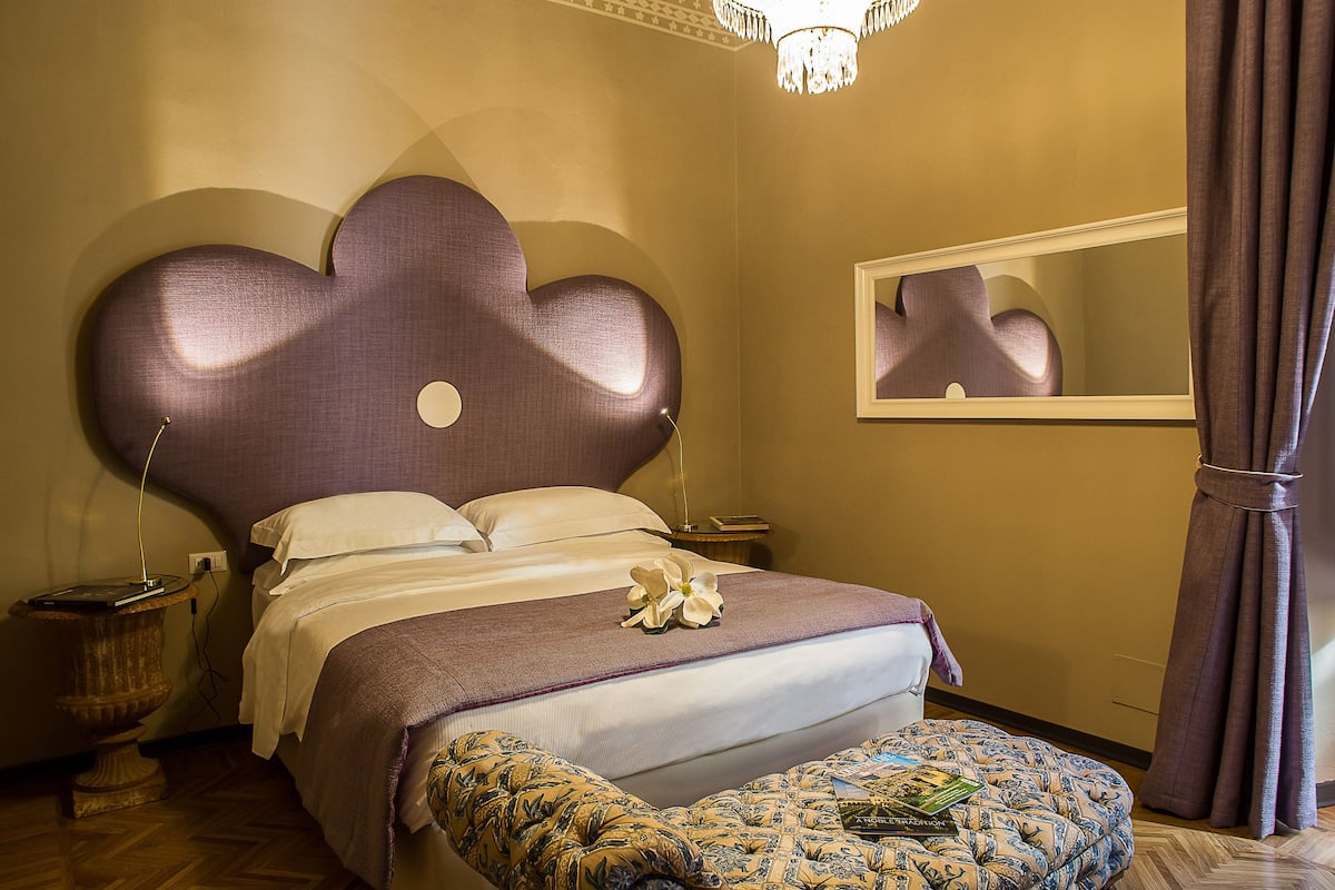 A bedroom with a large bed, a nightstand, a mirror, and a patterned ottoman.
