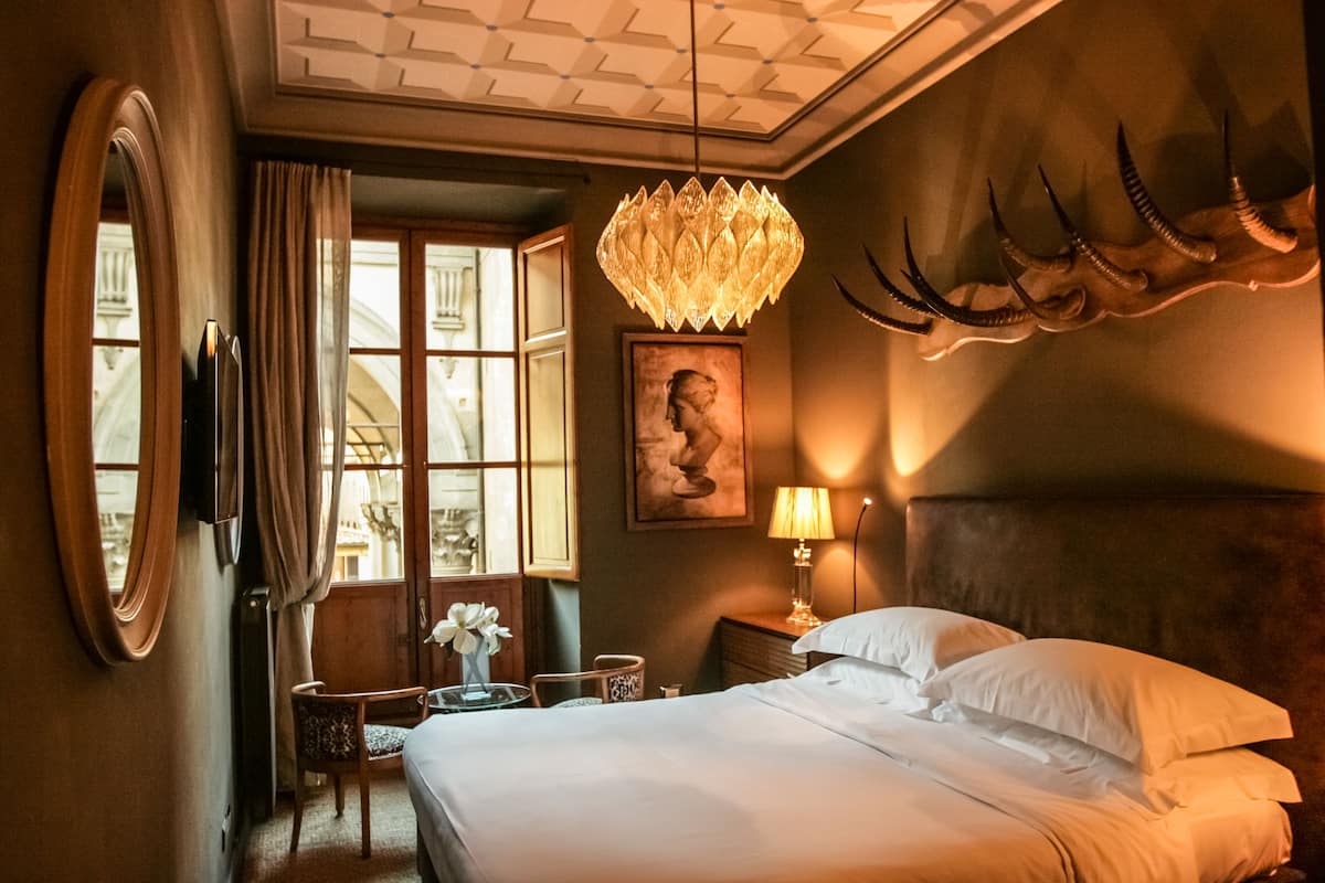 A hotel room with a bed, a chandelier, a mounted animal head, a painting, and a window with curtains.