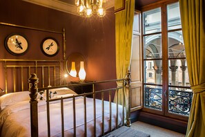 A bedroom with a bed, a chandelier, a window with a view of buildings, and a clock on the wall. - Porcellino Gallery (Florence)