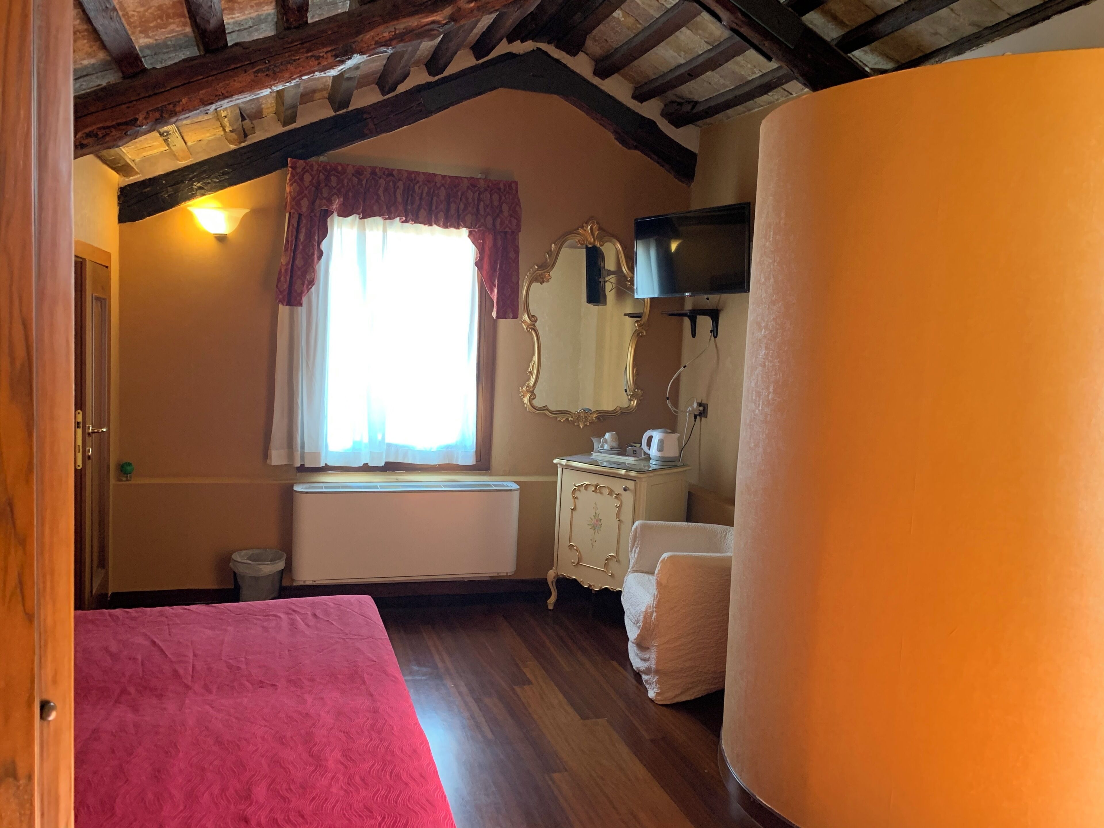 A bedroom with a wooden ceiling, a radiator, a window with curtains, a vanity with a mirror, and a bed with a pink bedspread.