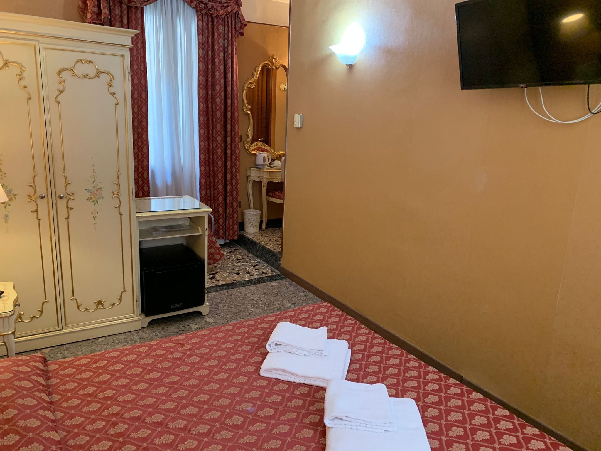 A hotel room with a bed, a wardrobe, a vanity with a mirror, and a television mounted on the wall.