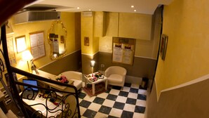 Ein Hotelzimmer mit Schachbrettmuster-Boden, einem Schreibtisch mit einem Laptop, einem Stuhl, einem kleinen Tisch mit Blumen und einem Spiegel an der Wand.