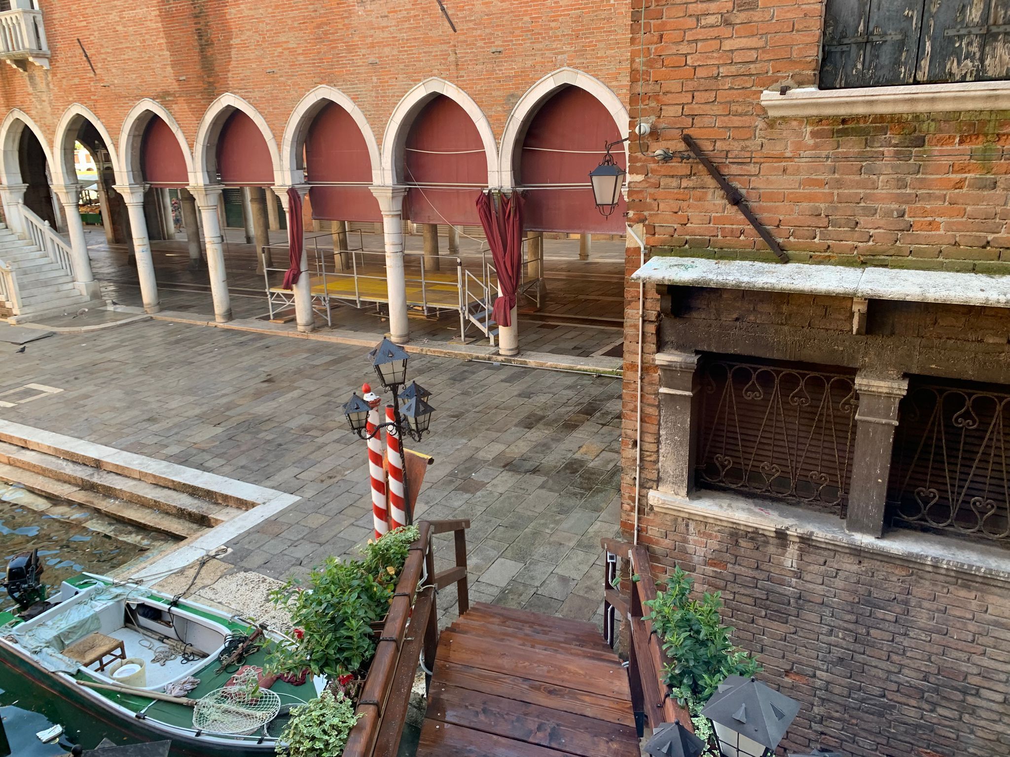 Ein Innenhof mit einem Backsteingebäude, das rundbogige Durchgänge, ein am Ufer vertäutes Boot und eine kleine Holzbank zeigt.