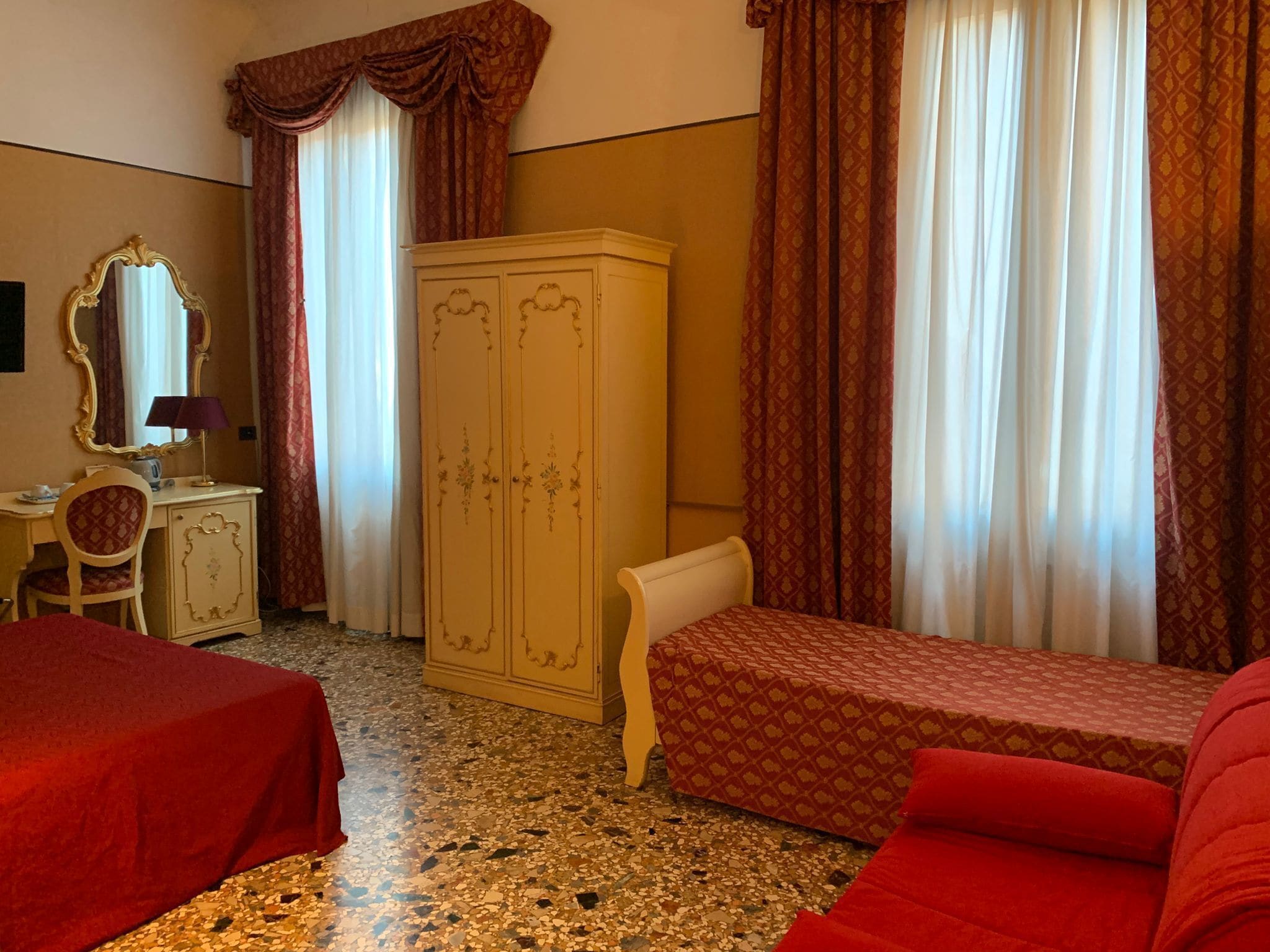 Une chambre d’hôtel avec deux lits simples, une armoire, un bureau avec une chaise, un miroir et une fenêtre avec des rideaux.