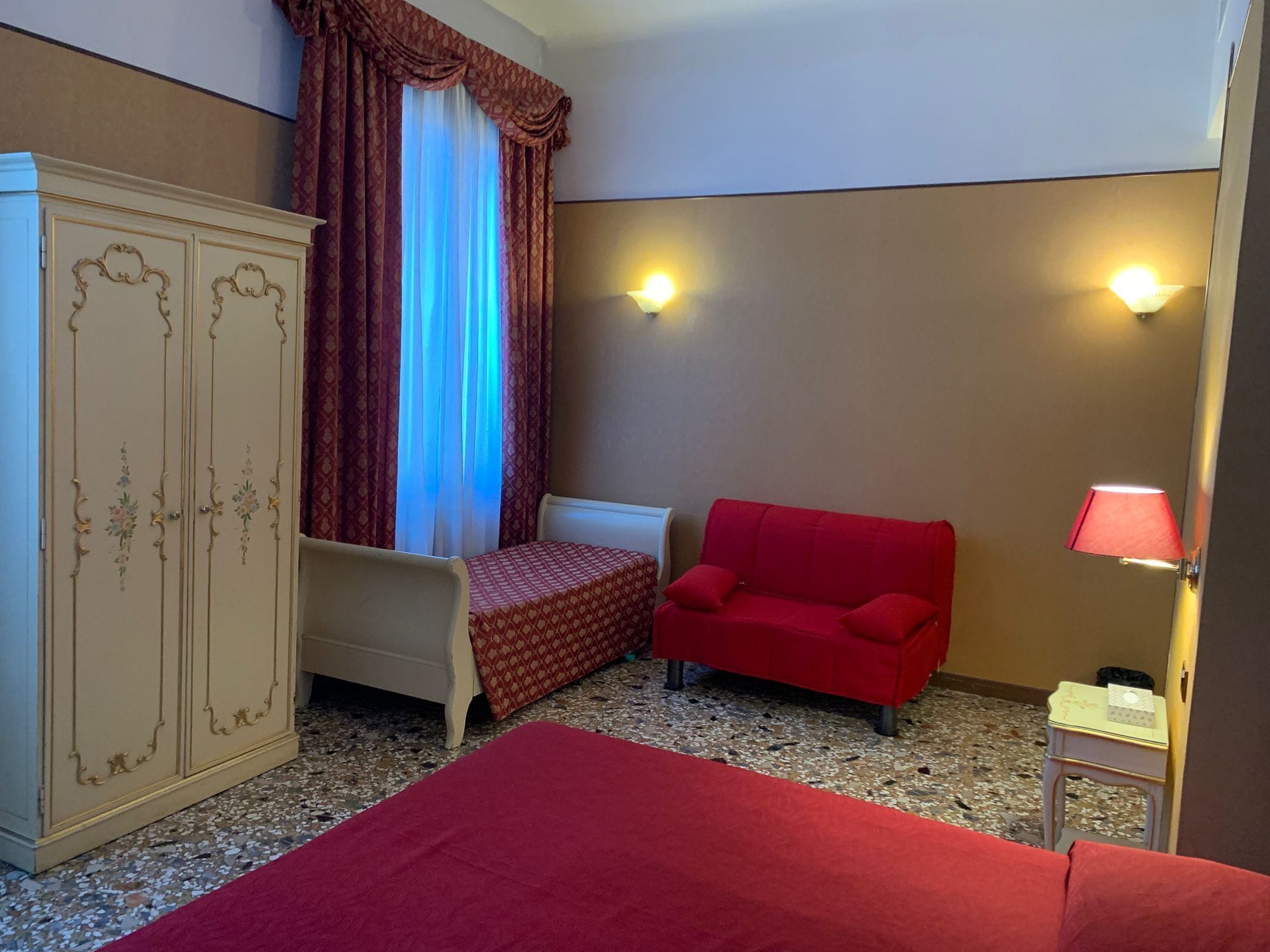 A hotel room with a bed, a red sofa, a small table with a lamp, a wardrobe, and a window with curtains.