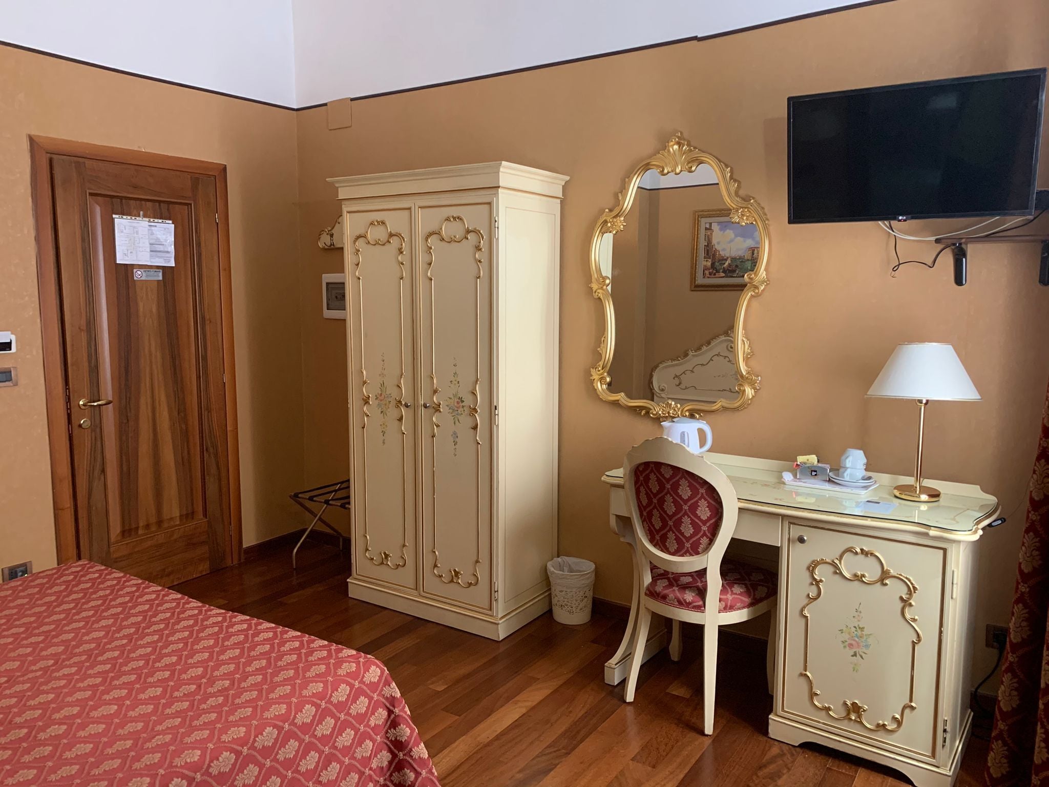 A hotel room with a wooden wardrobe, a desk with a chair, a television, and a bed with a patterned bedspread.