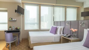 Habitación de hotel con dos camas, un escritorio con silla, televisor montado en la pared y vista a la ciudad a través de amplias ventanas.