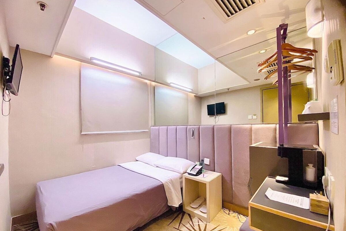 Habitación de hotel con una cama individual, una mesita de noche pequeña, un espejo, un televisor y una ventana con persianas.