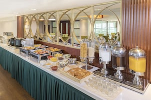 Buffet com diversos alimentos e bebidas, um espelho com moldura decorativa e uma área de estar ao fundo.