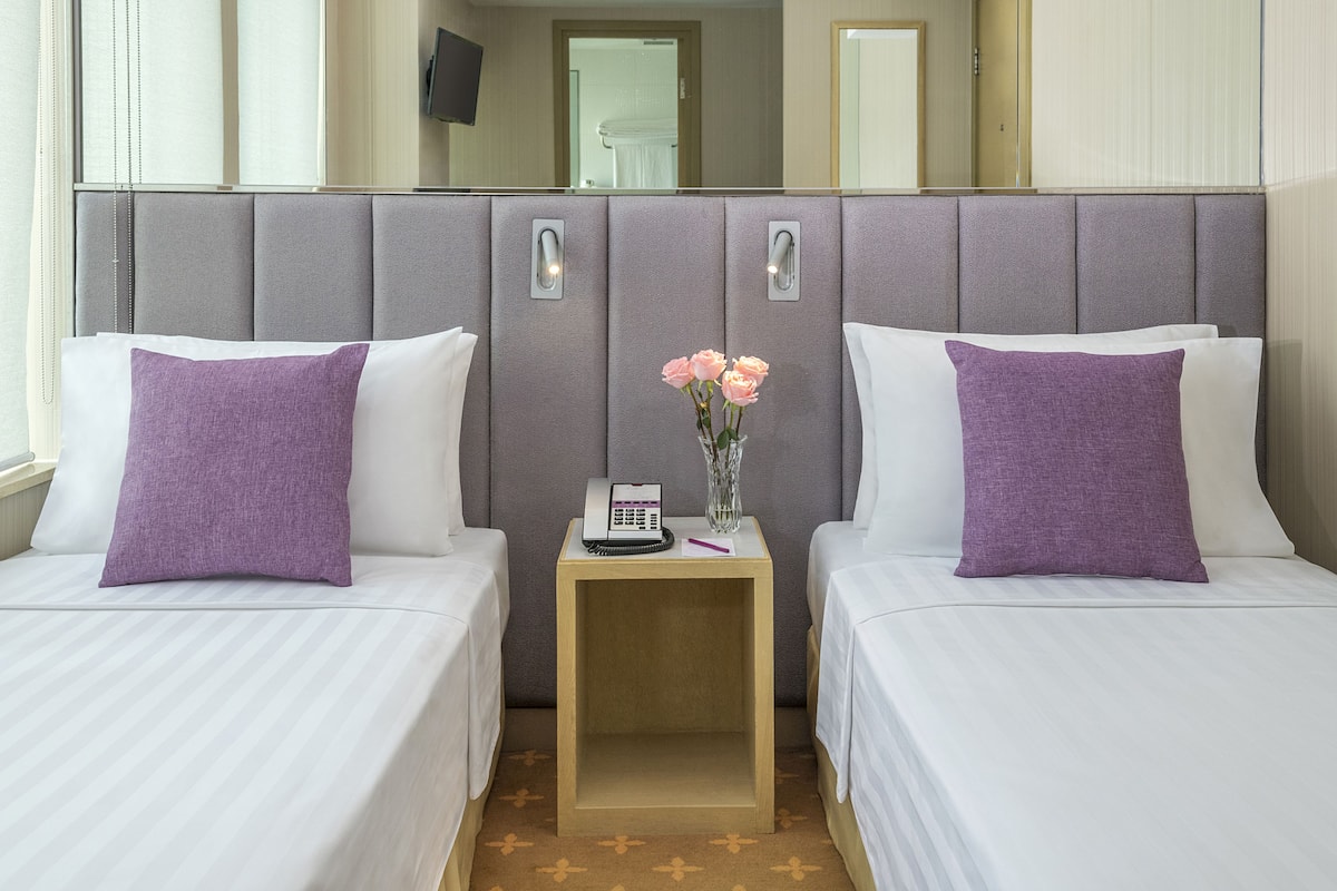 Une chambre d’hôtel avec deux lits, une table de chevet avec un téléphone et un vase, et un miroir au-dessus des lits.