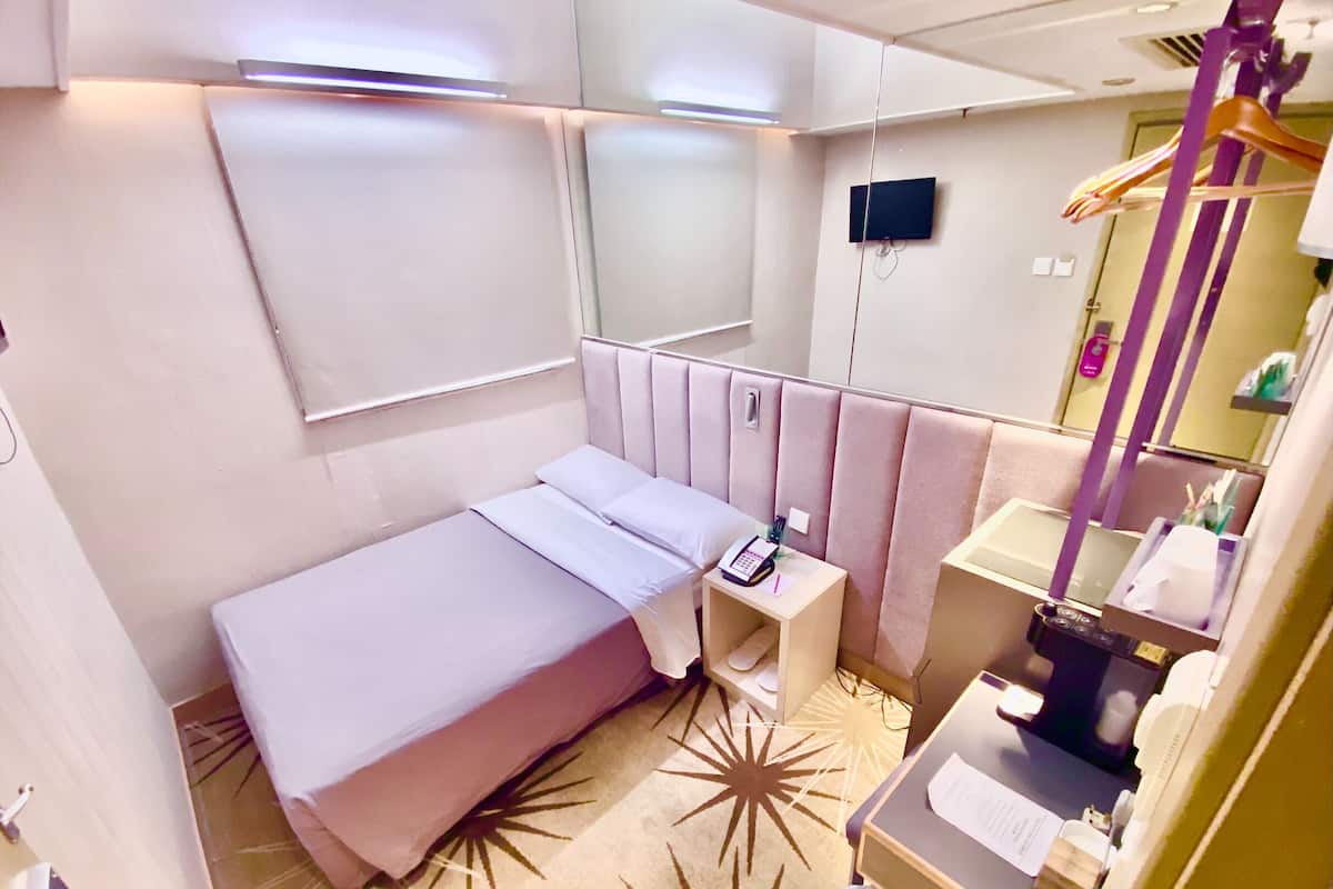 Une chambre d’hôtel compacte comprenant un lit simple, une table de chevet avec un téléphone, un petit bureau avec un ordinateur, un miroir et une salle de bain avec un lavabo et un porte-serviettes.