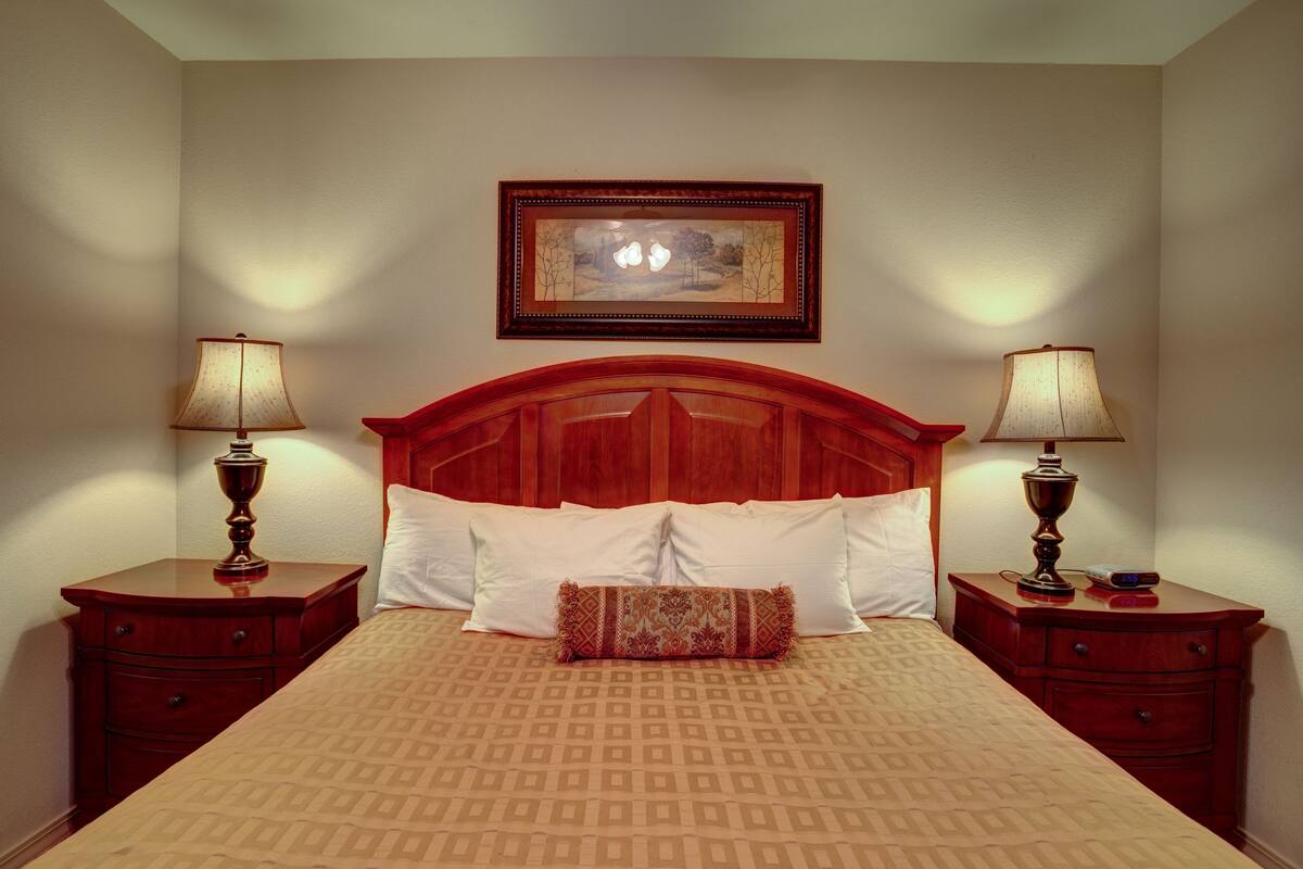 Una habitación de hotel con cabecera de madera, dos mesitas de noche con lámparas, una cama con colcha estampada y un cuadro en la pared.