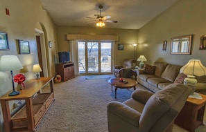 A living room with a sofa, coffee table, and a television on a stand. There is a ceiling fan, a lamp, and a window with blinds.