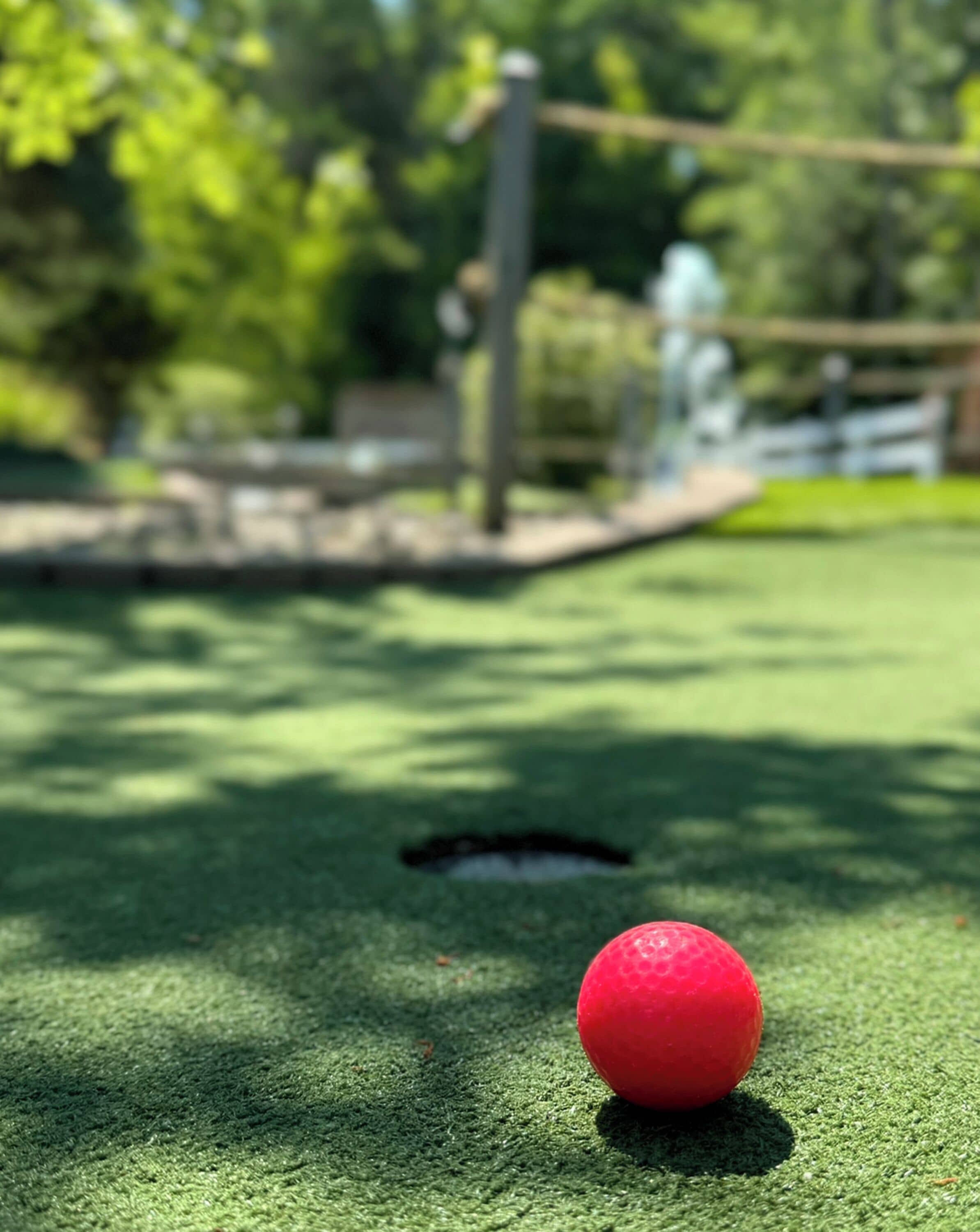 Ein roter Golfball auf einem grünen Kunstrasenplatz, im Hintergrund ein Loch.