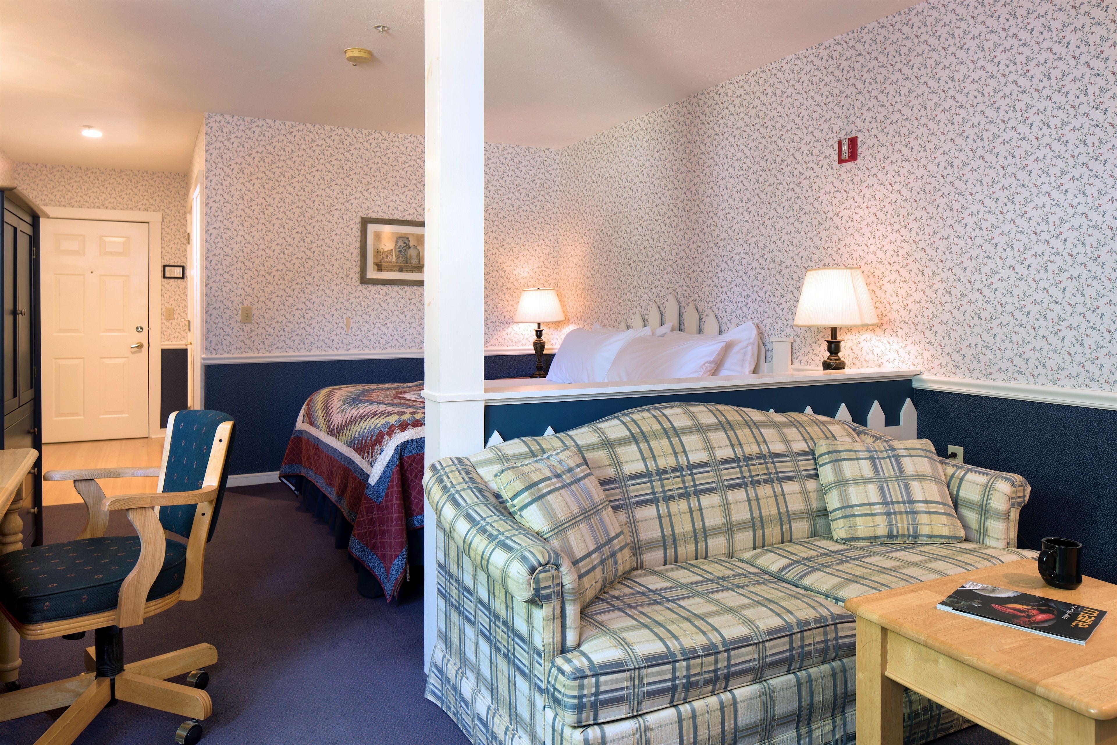 A room with a plaid sofa, a wooden chair, a bed with a quilt, two bedside lamps, and a small table with a book.