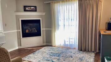 Ein Wohnzimmer mit Kamin, einem großen Teppich und Blick ins Freie durch ein Fenster mit Vorhängen.