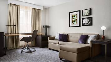 A modern hotel room with a beige sofa, a desk, and a chair. There are two framed artworks on the wall and a lamp on the side table.