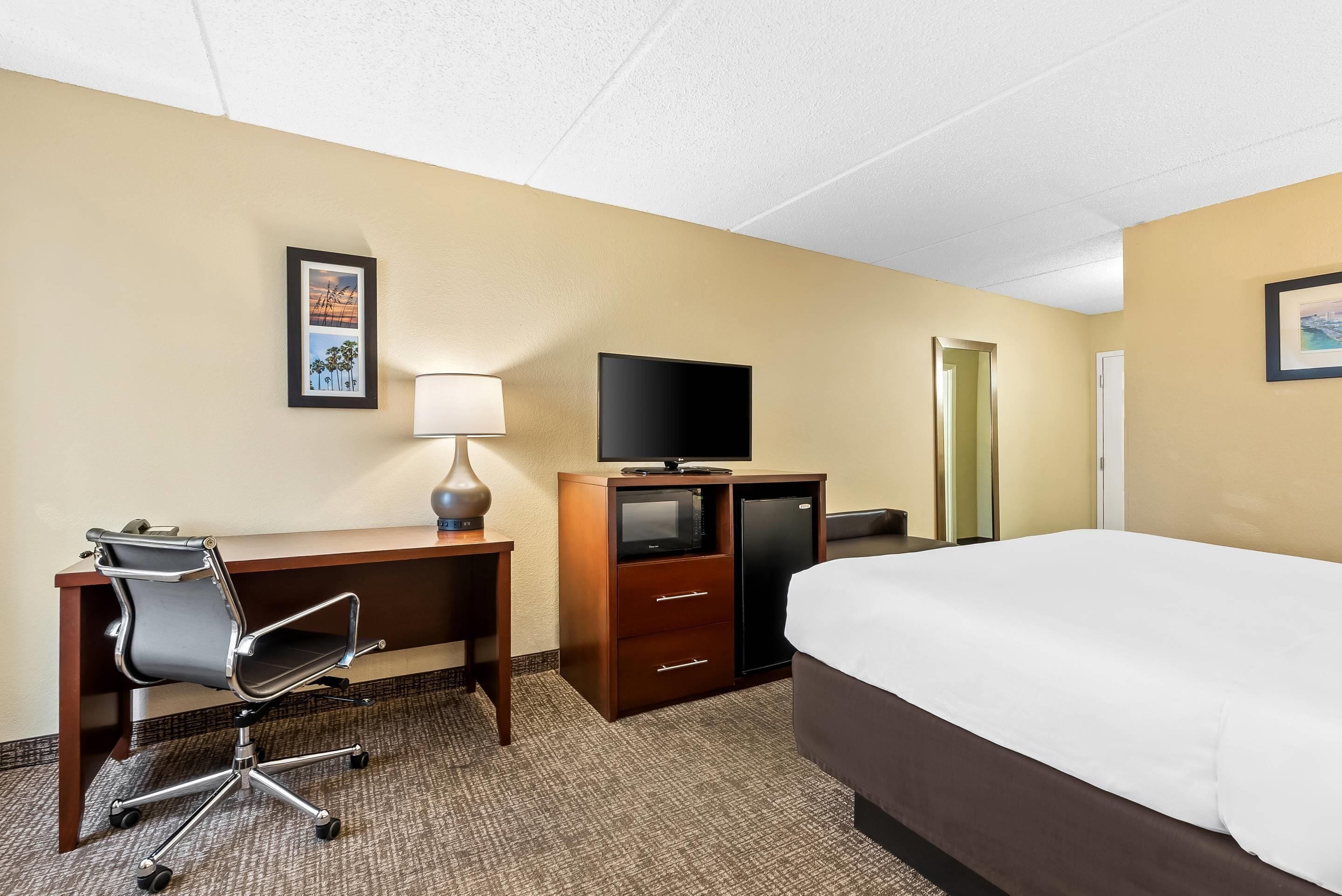 Quarto de hotel com cama, escrivaninha com cadeira, televisão, cômoda e espelho.