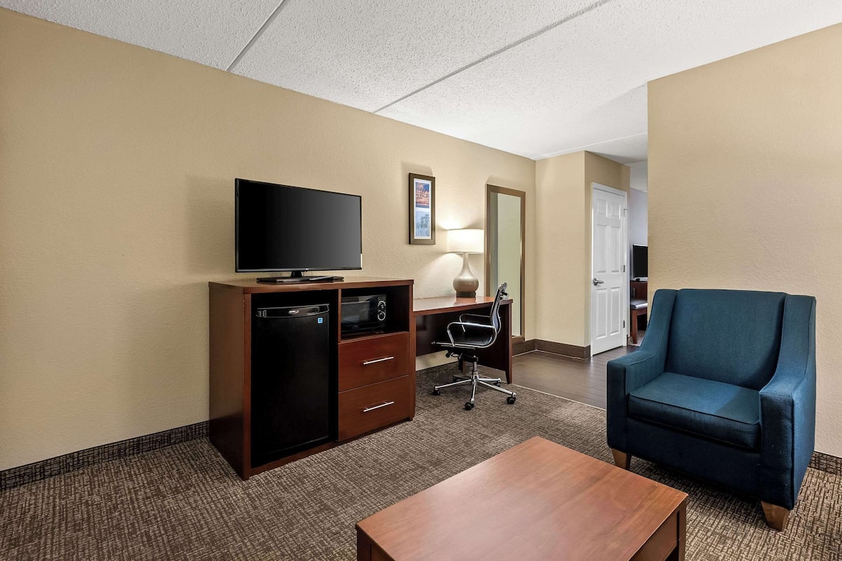 A hotel room with a flat-screen TV on a wooden cabinet, a desk with a chair, a blue armchair, and a small wooden table.