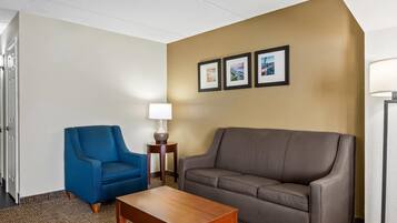Une chambre d’hôtel avec un fauteuil bleu, un canapé marron, une table basse en bois et une lampe. Trois tableaux encadrés sont accrochés au mur.