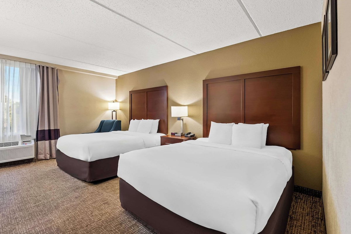 A hotel room with two beds, a large window with curtains, a wall-mounted TV, and a bedside table with a lamp.