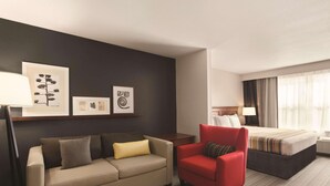 A hotel room with a sofa, a red armchair, a bed, and artwork on the wall. - Country Inn & Suites by Radisson, Bentonville South - Rogers, AR (Rogers)