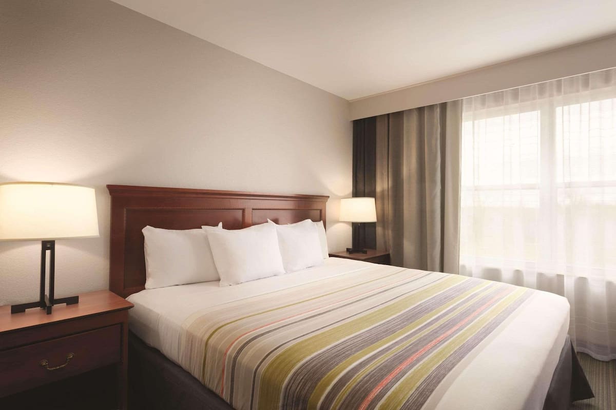A hotel room with a large bed, two bedside tables with lamps, a wooden headboard, and sheer curtains on the window.