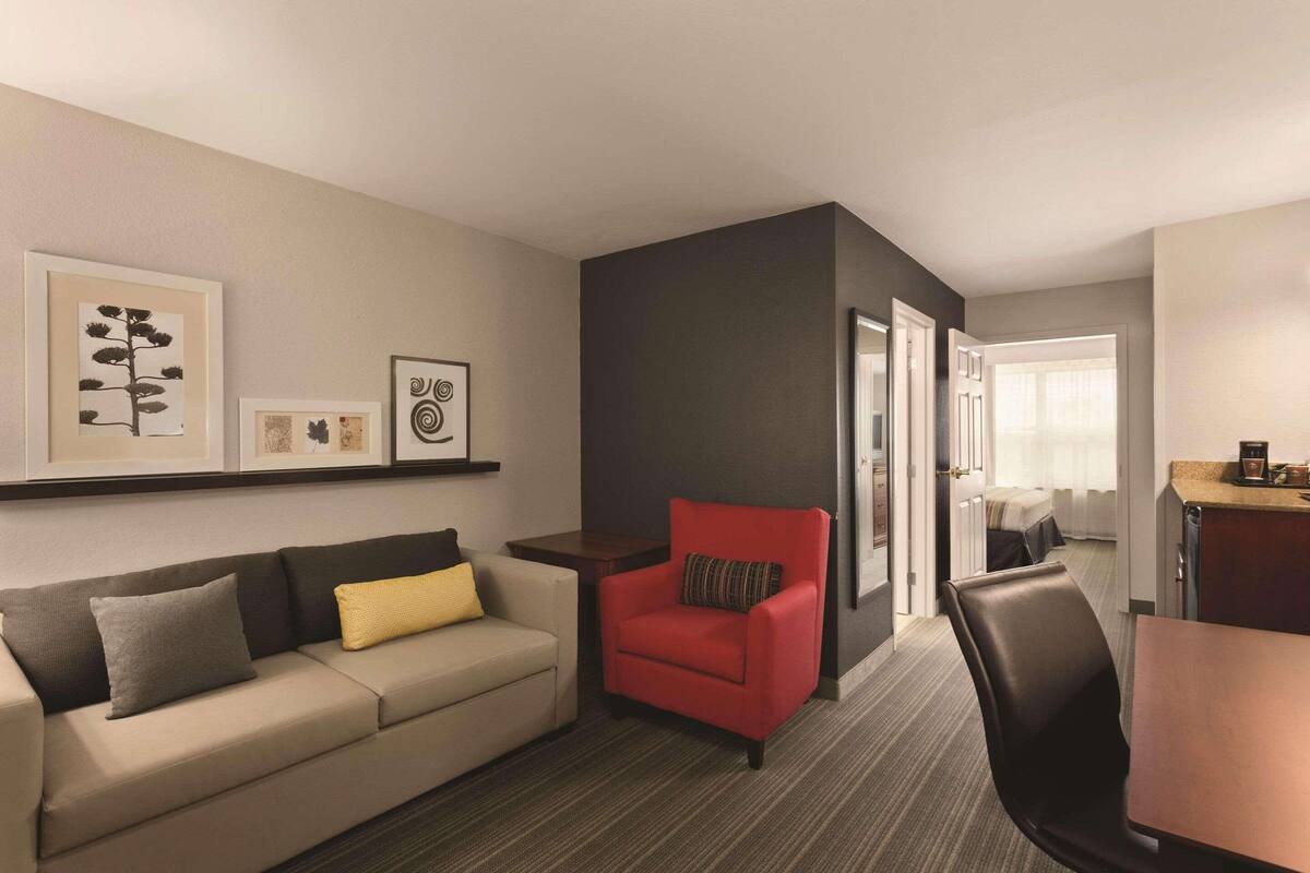 A hotel room with a sofa, a red armchair, a dining table, and a kitchen area.