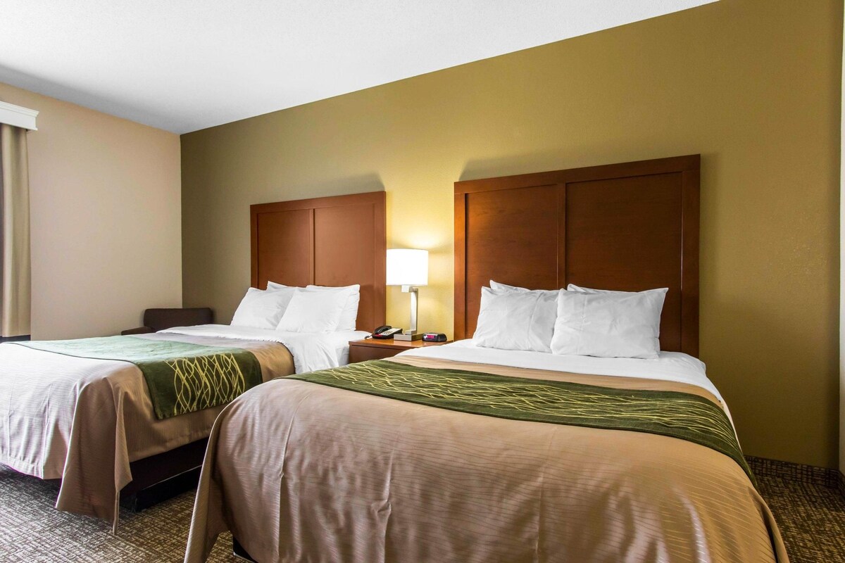 A hotel room with two beds, each with a wooden headboard, a bedside table with a lamp, and a green throw blanket.
