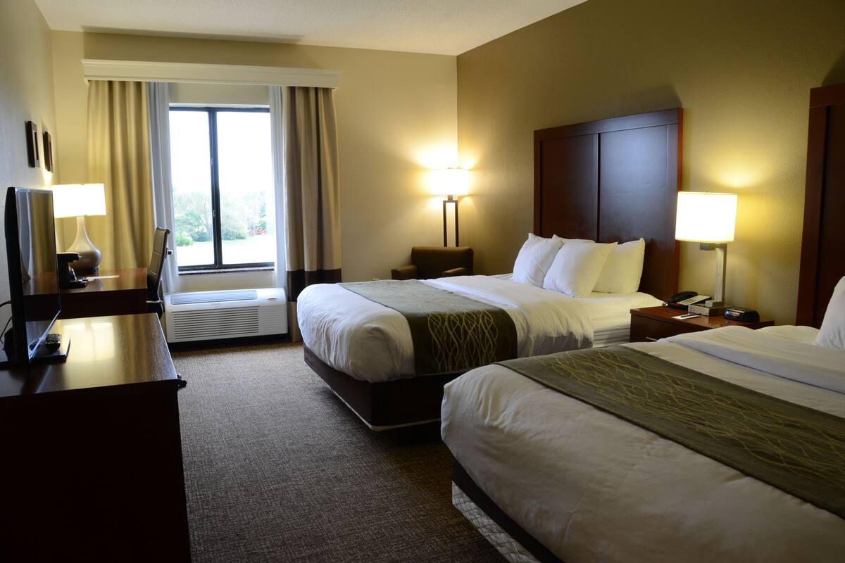 A hotel room with two beds, a desk with a lamp, a chair, a nightstand with a phone, and a window with curtains.