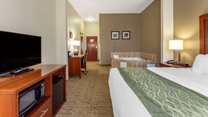 A hotel room with a bed, a TV, a desk with a lamp, a microwave, a bathroom with a bathtub, and a door leading to another room.