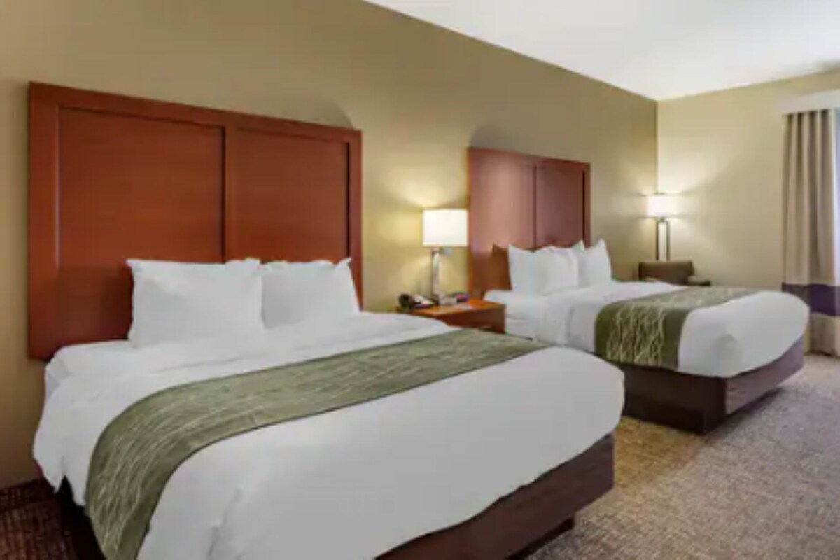 A hotel room with two beds, a wooden headboard, a nightstand with a lamp, and a window with curtains.