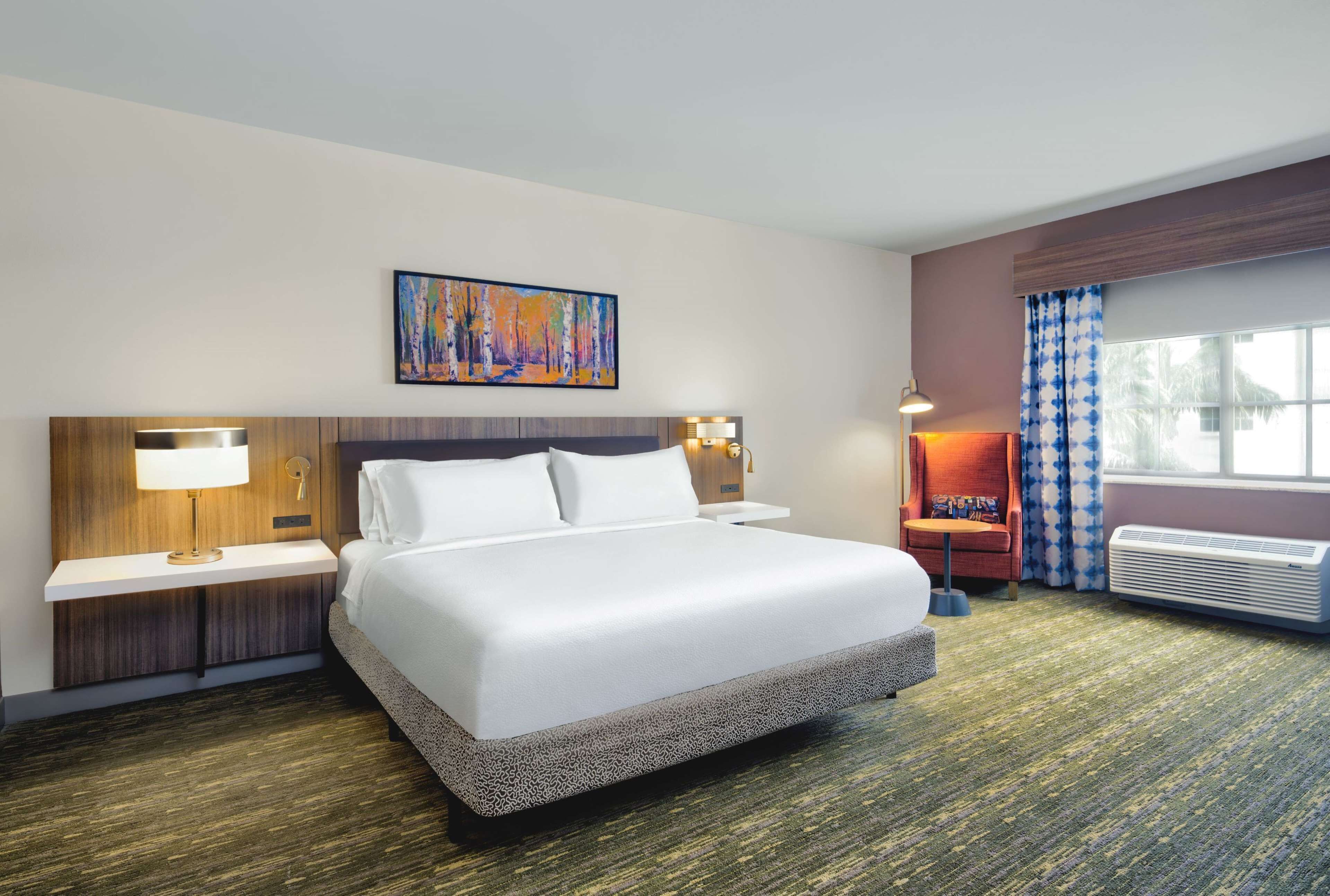 Habitación de hotel con una cama grande, una mesita de noche con lámpara, una obra de arte en la pared, una ventana con cortinas y un equipo de aire acondicionado.
