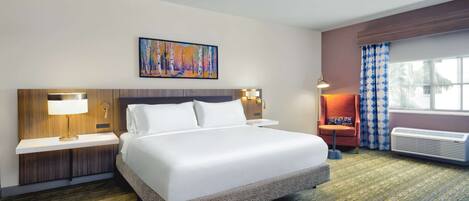 Habitación de hotel con una cama grande, una mesita de noche con lámpara, una obra de arte en la pared, una ventana con cortinas y un equipo de aire acondicionado.