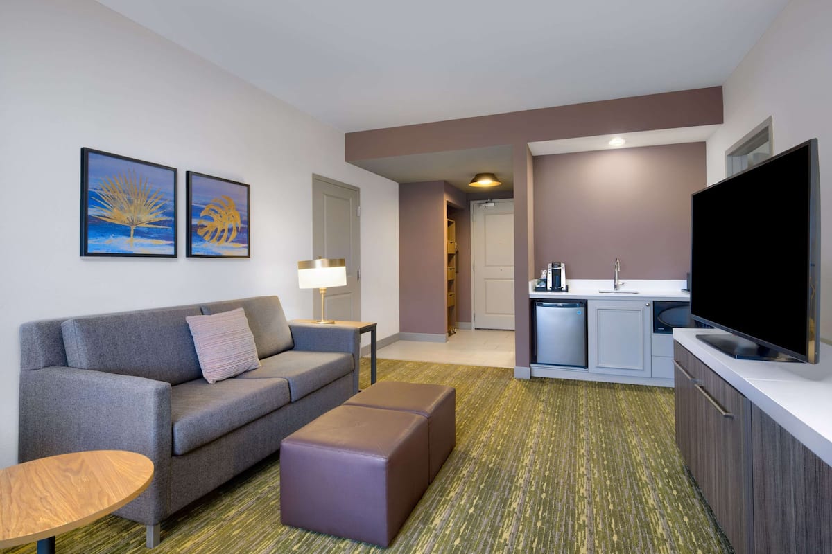 A modern hotel room with a grey sofa, a small wooden table, a purple ottoman, a flat-screen TV, and a kitchenette with a sink and stove.