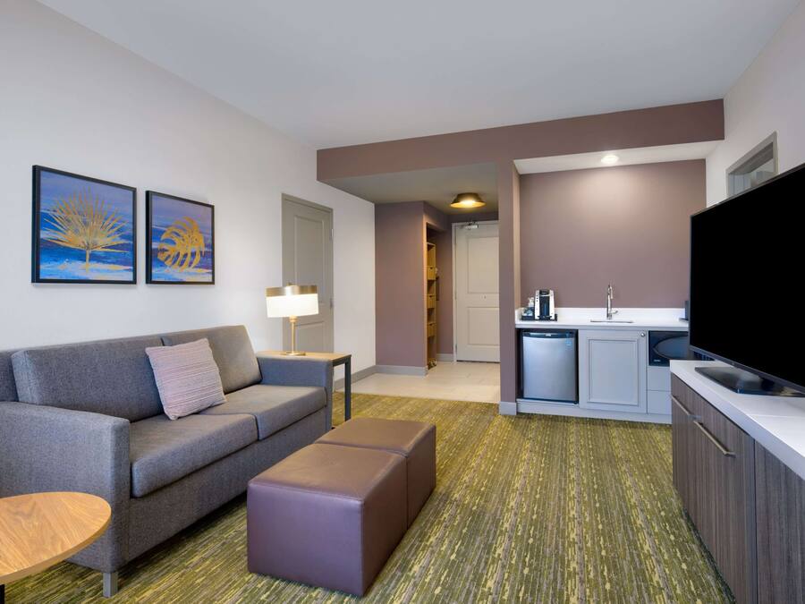Una habitación de hotel moderna con un sofá gris, una pequeña mesa de madera, un reposapiés morado, un televisor de pantalla plana y una kitchenette con un lavabo y estufa.