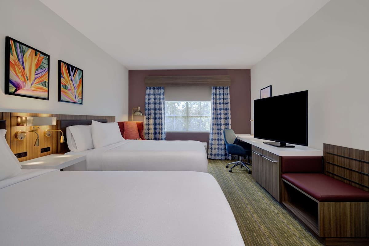 A hotel room with two beds, a desk with a television, a window with curtains, and artwork on the wall.