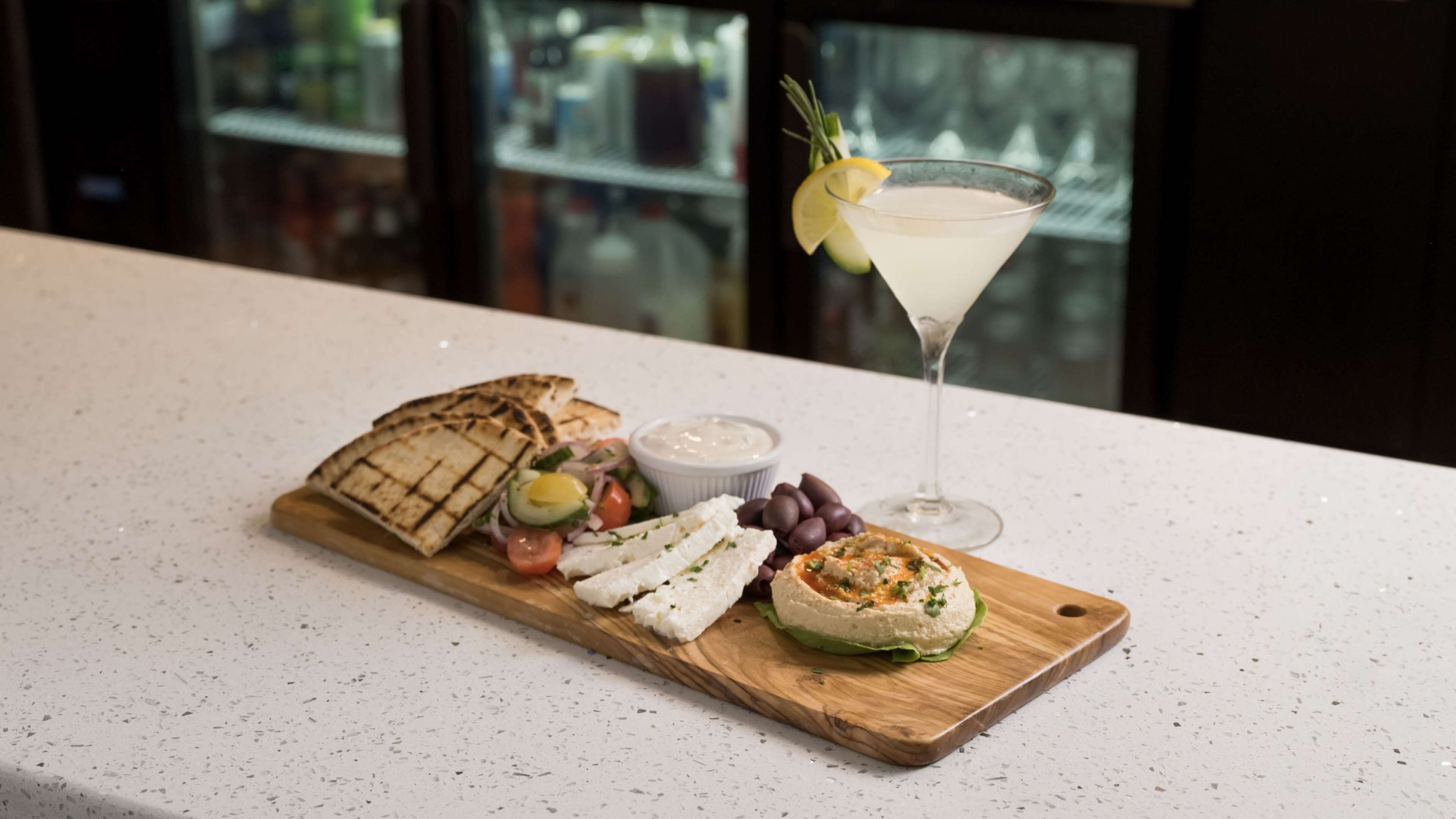 A wooden board with grilled cheese, olives, a salad, and a martini on a countertop.