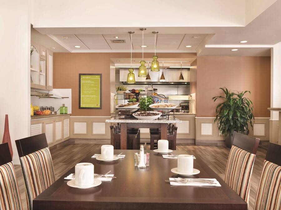 A dining area with a long table set for a meal, chairs with striped upholstery, and a kitchen in the background.