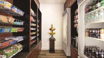A narrow hallway with shelves stocked with snacks, a refrigerator with bottled water, and a small table with fruit.