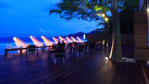 An outdoor dining area with wooden flooring, tables, and chairs, overlooking a body of water and mountains in the background. - The Library (Koh Samui)