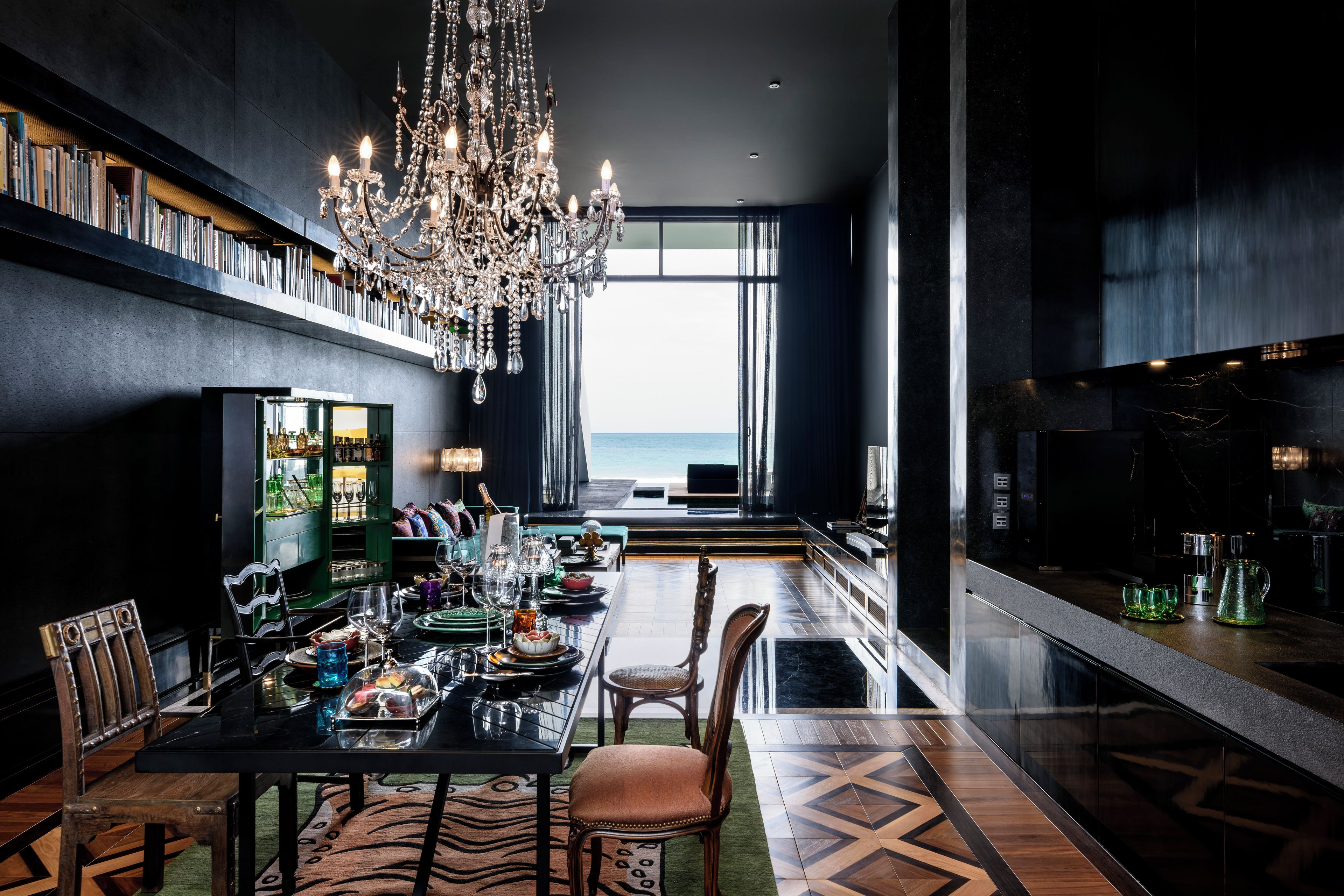 A modern dining area with a chandelier, a view to the sea, and a bar area with a marble countertop.