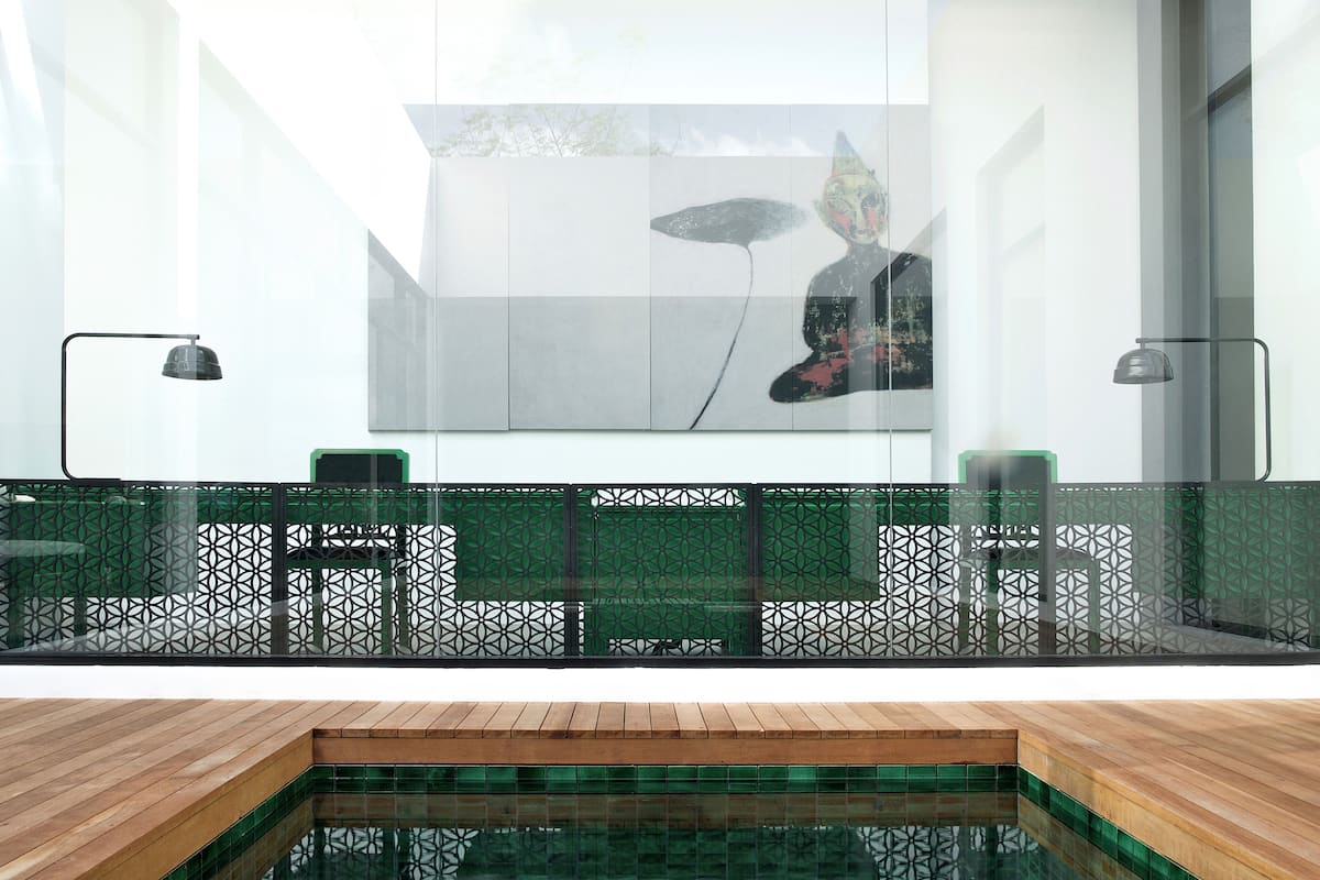 A modern indoor pool area with a wooden deck, a glass partition with a geometric pattern, and a large abstract painting on the wall.
